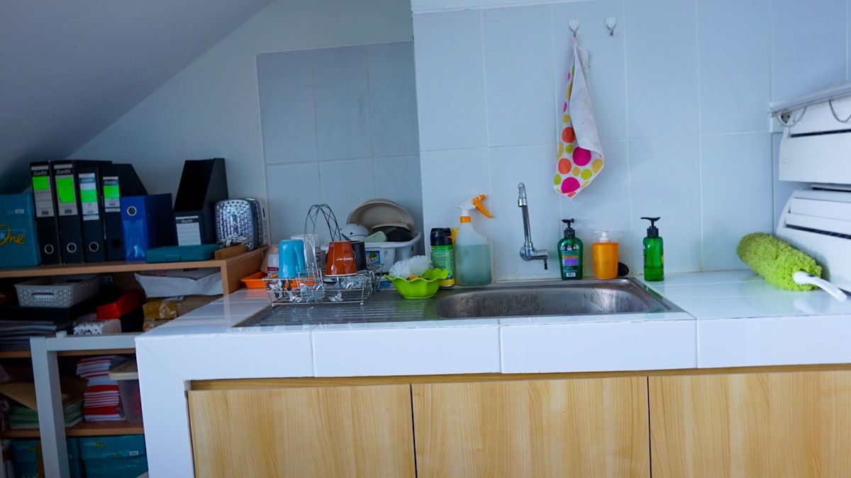 A cramped apartment kitchen with a sink, stove, and refrigerator set against shared walls, showing roaches moving along the baseboard, around a pipe opening under the sink, and near a small gap behind the appliance, with one roach on a light switch in the foreground