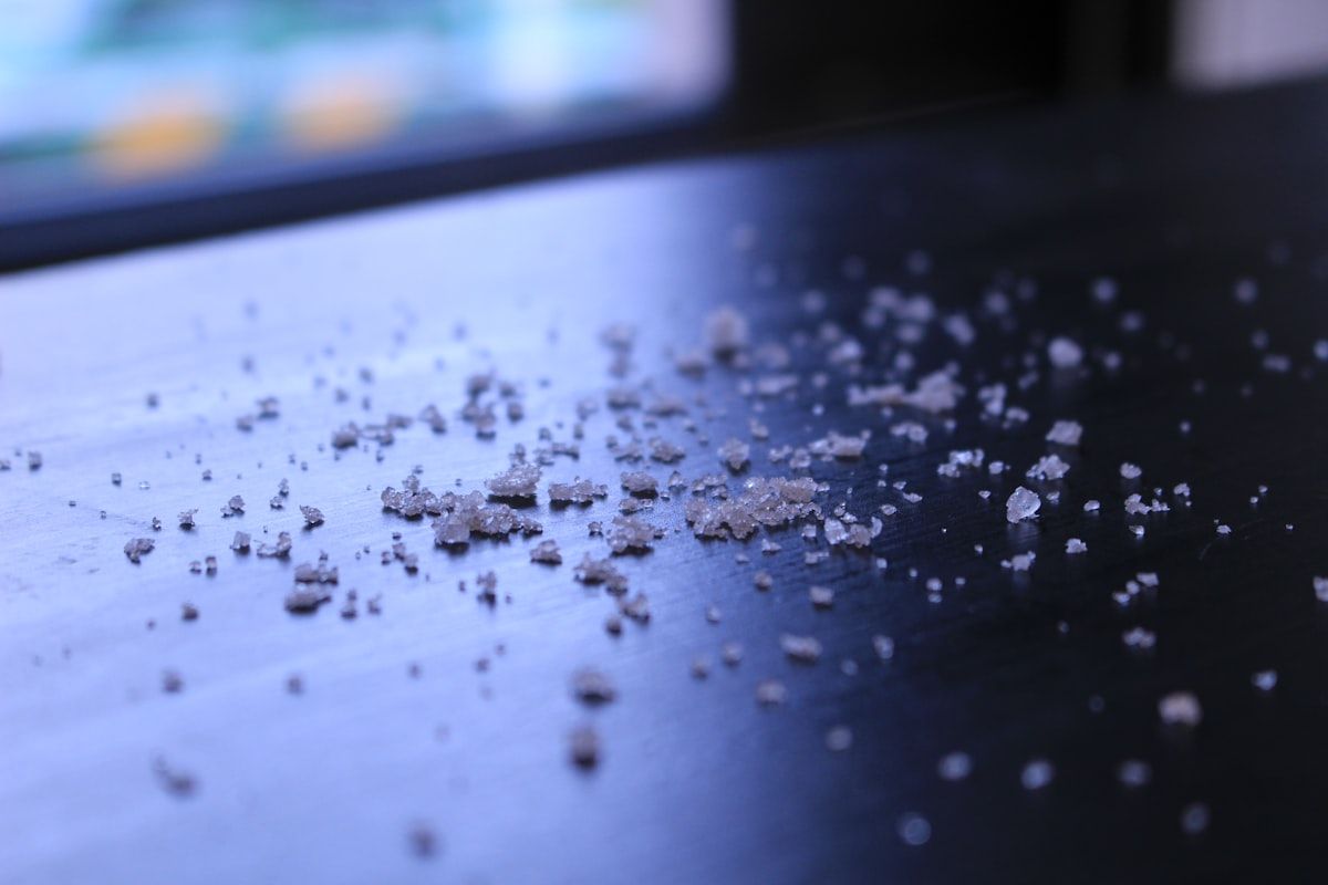 A close-up view of several cockroach egg cases on the edge of a cabinet shelf near crumbs and a damp kitchen corner, with tiny dark roach nymphs clustered nearby to illustrate rapid indoor breeding and growing infestation.