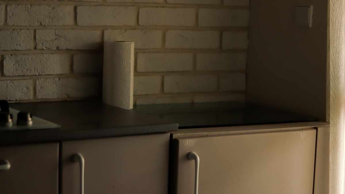 A close-up of a kitchen wall gap behind a dishwasher and cabinet toe kick, showing bait stations, a dust applicator near a crack in the wall, and sticky monitoring traps placed along the floor where roaches would travel