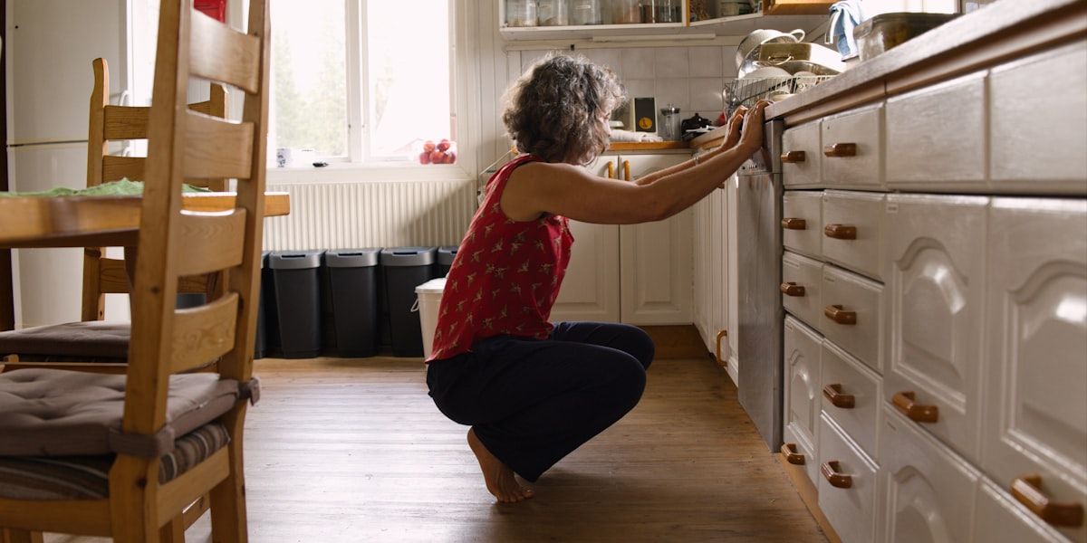 A pest control technician kneeling beside a kitchen cabinet opening, placing gel bait into hinges and cracks with a small applicator while dust is being applied into a wall gap near plumbing pipes; a spray bottle, sticky monitor traps, and cabinet doors are open around the work area.