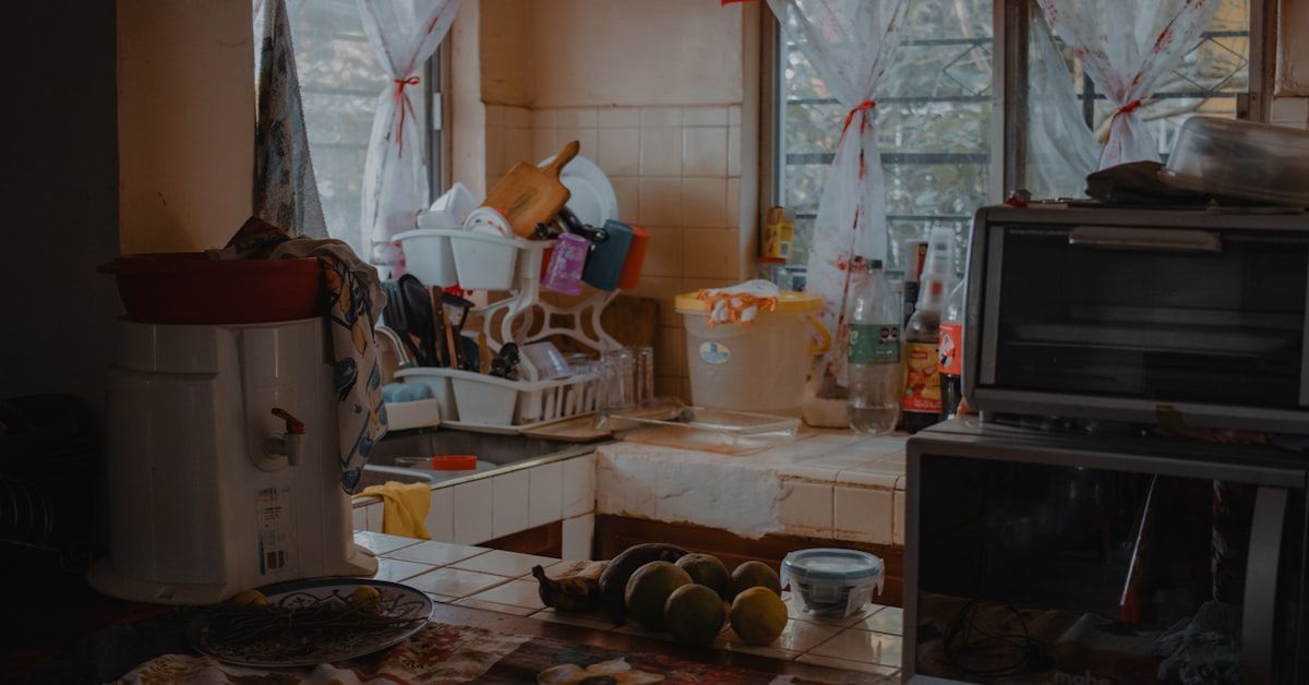 A kitchen cleanup scene with sealed food containers on the counter, an open trash can being emptied, cardboard boxes stacked near the door ready to leave, and a person wiping crumbs from the stove area while pulling clutter away from the wall under the sink